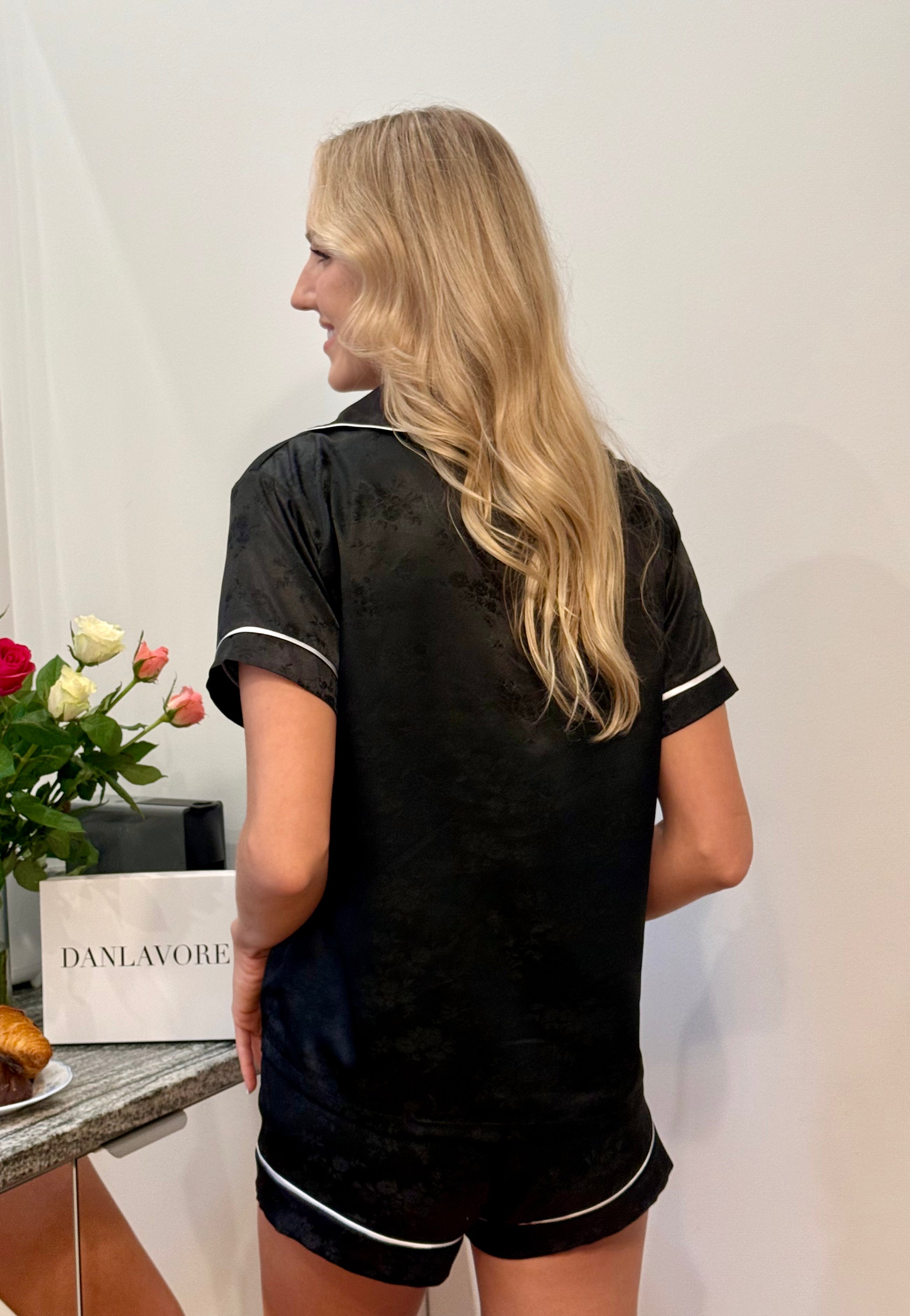 Woman wearing a black outfit with white trim, standing in front of a white wall with a 'DANLAVORE' box and flowers on a table.