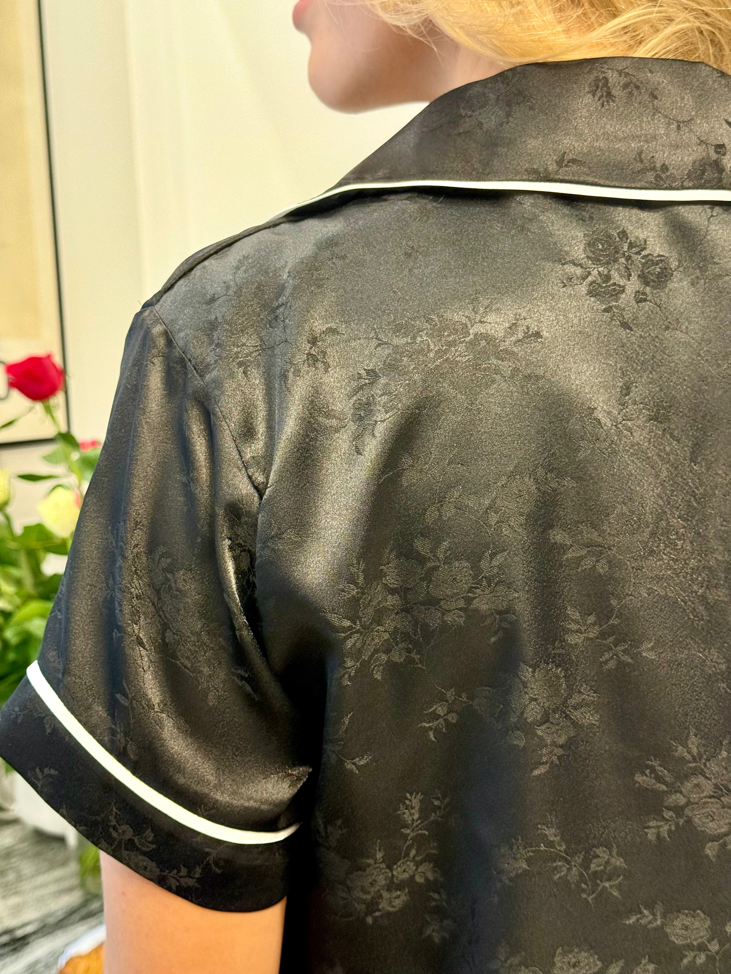 Person wearing a black floral satin robe with white trim, standing in front of a blurred background.